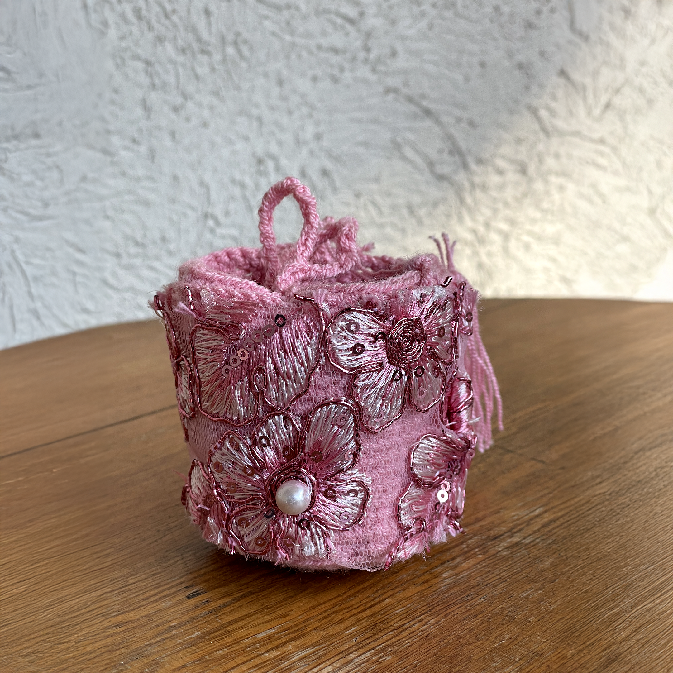 Mini mochila Wayúu color rosa con bordados florales en relieve, lentejuelas y detalle de perla central sobre mesa de madera.