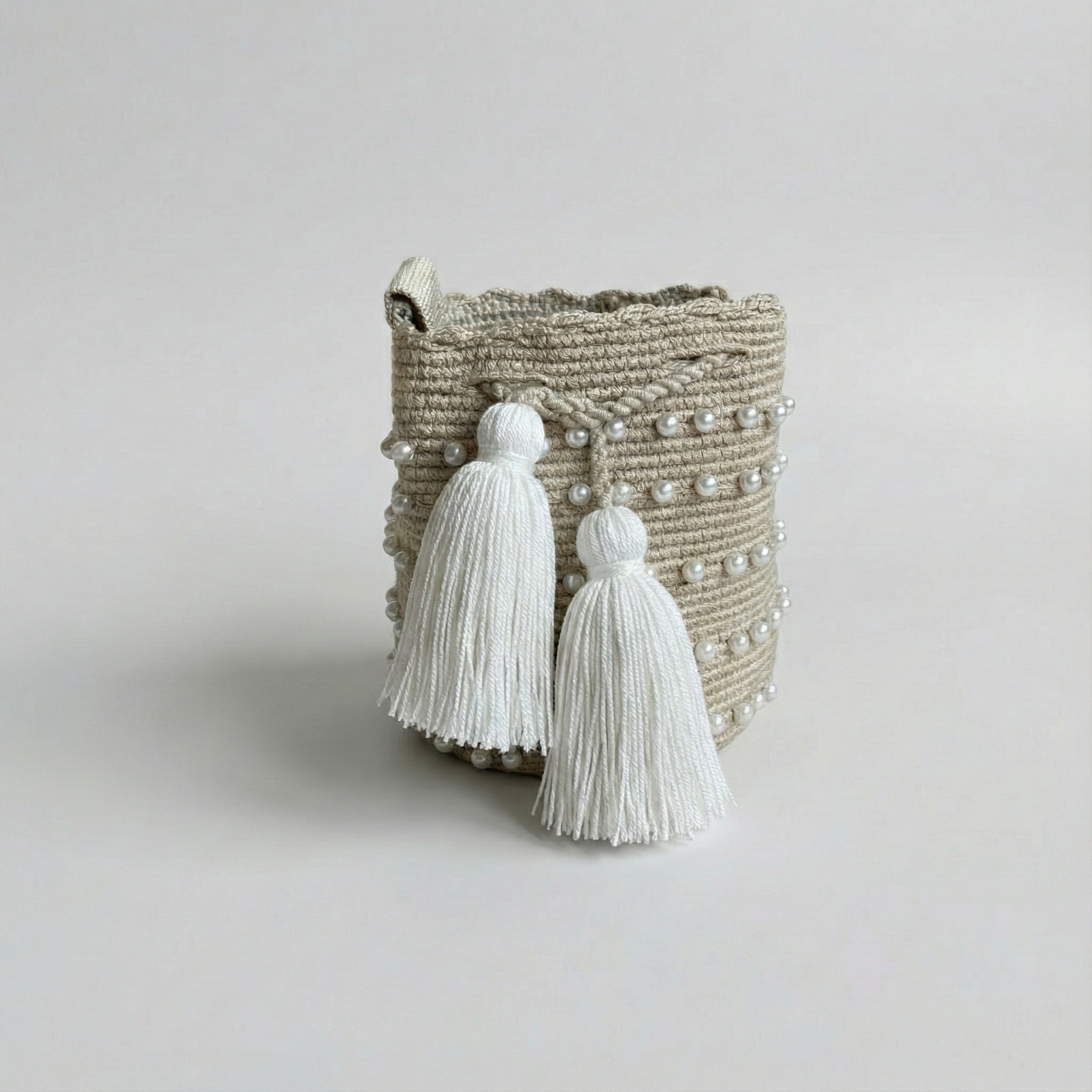 Fotografía de producto con fondo blanco de una mochila pequeña tejida a crochet en color beige, decorada con hileras de perlas blancas incrustadas y dos borlas grandes de hilo blanco colgando del cierre frontal