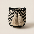 Mochila Wayuu artesanal tejida a mano con patrón de zigzag en colores negro y beige. Diseño de balde con cordón de cierre y dos borlas decorativas grandes de color claro sobre una mesa de madera.