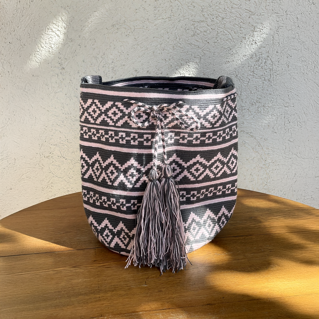 Mochila Wayúu tejida con patrones geométricos en rosado claro y gris, con cierre de cordón y borlas grandes.
