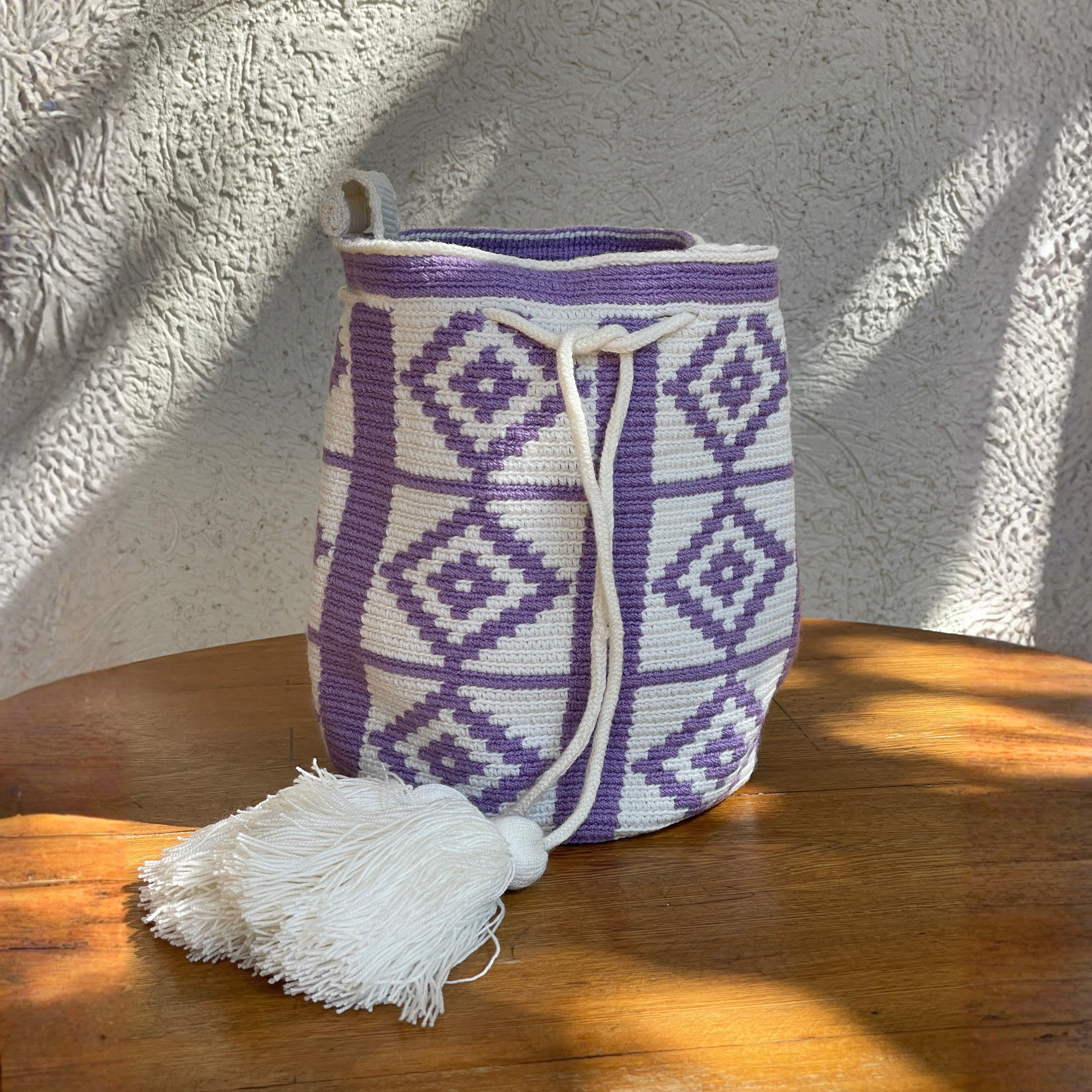 Mochila Wayúu artesanal con diseño de rombos geométricos en color lila y blanco, cordón de cierre blanco y una borla grande de flecos sobre una mesa de madera.