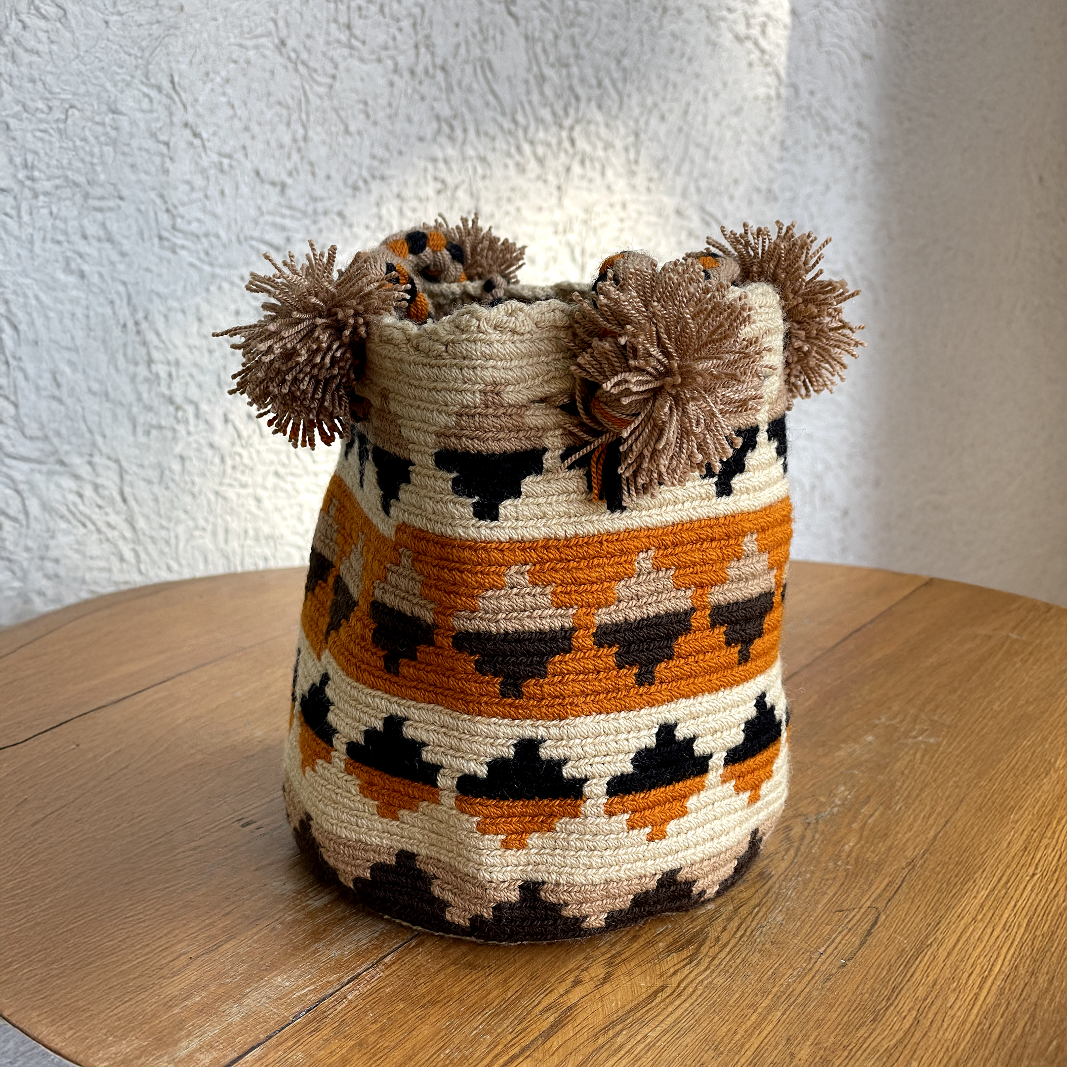 Mochila Wayúu tejida a mano con patrones geométricos en colores terracota, café y negro, decorada con borlas de hilo beige.