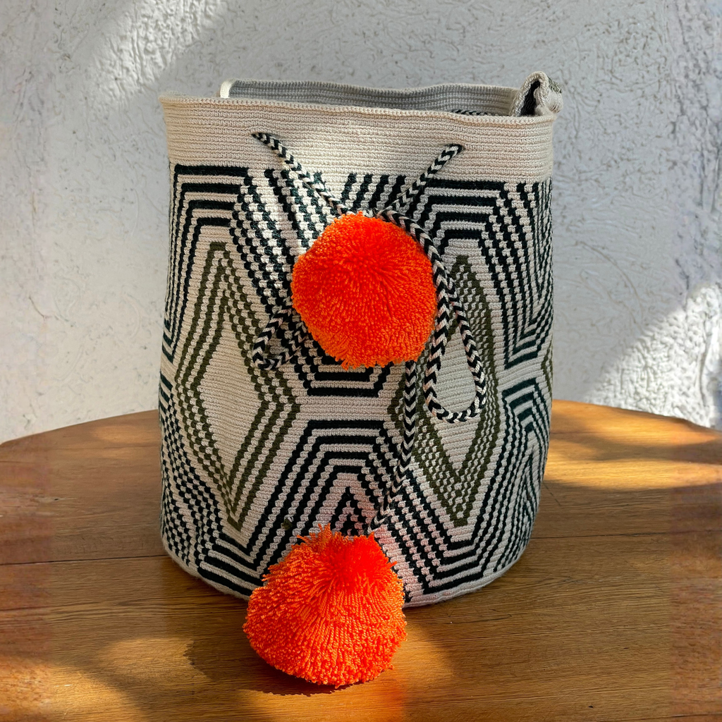Mochila Wayúu tejida a mano con patrones geométricos en color oliva, negro y crema, destacando dos pompones grandes color naranja brillante.