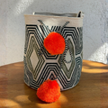 Mochila Wayúu tejida a mano con patrones geométricos en color oliva, negro y crema, destacando dos pompones grandes color naranja brillante.