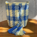 Mochila Wayúu tejida a mano con diseño de figuras geométricas en azul y crema, detalles de borlas grandes y cordón trenzado.