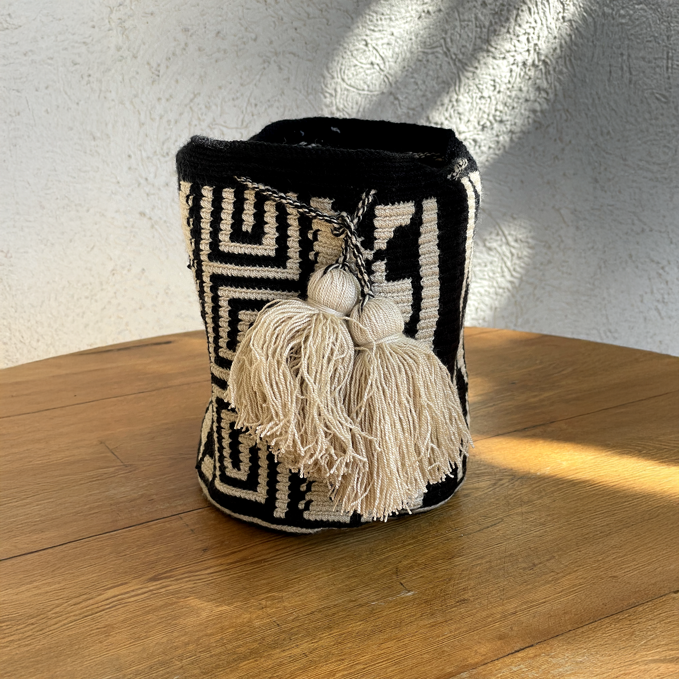 Mochila Wayúu con patrones geométricos Kanaas en blanco y negro, con dos borlas blancas grandes decorativas y cierre de cordón.