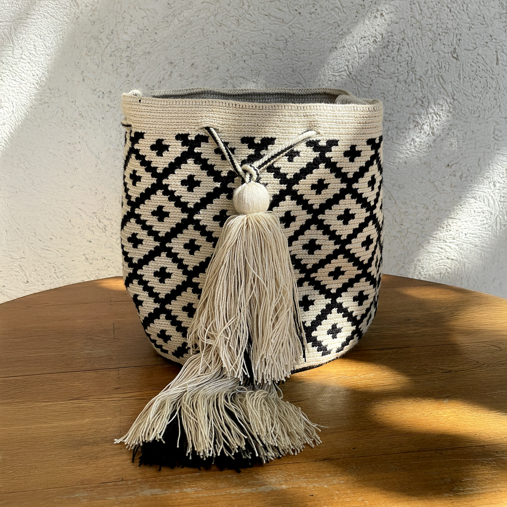 Mochila Wayúu con diseño de rombos negros sobre fondo beige, tejida a mano con técnica de crochet y decorada con una borla grande.