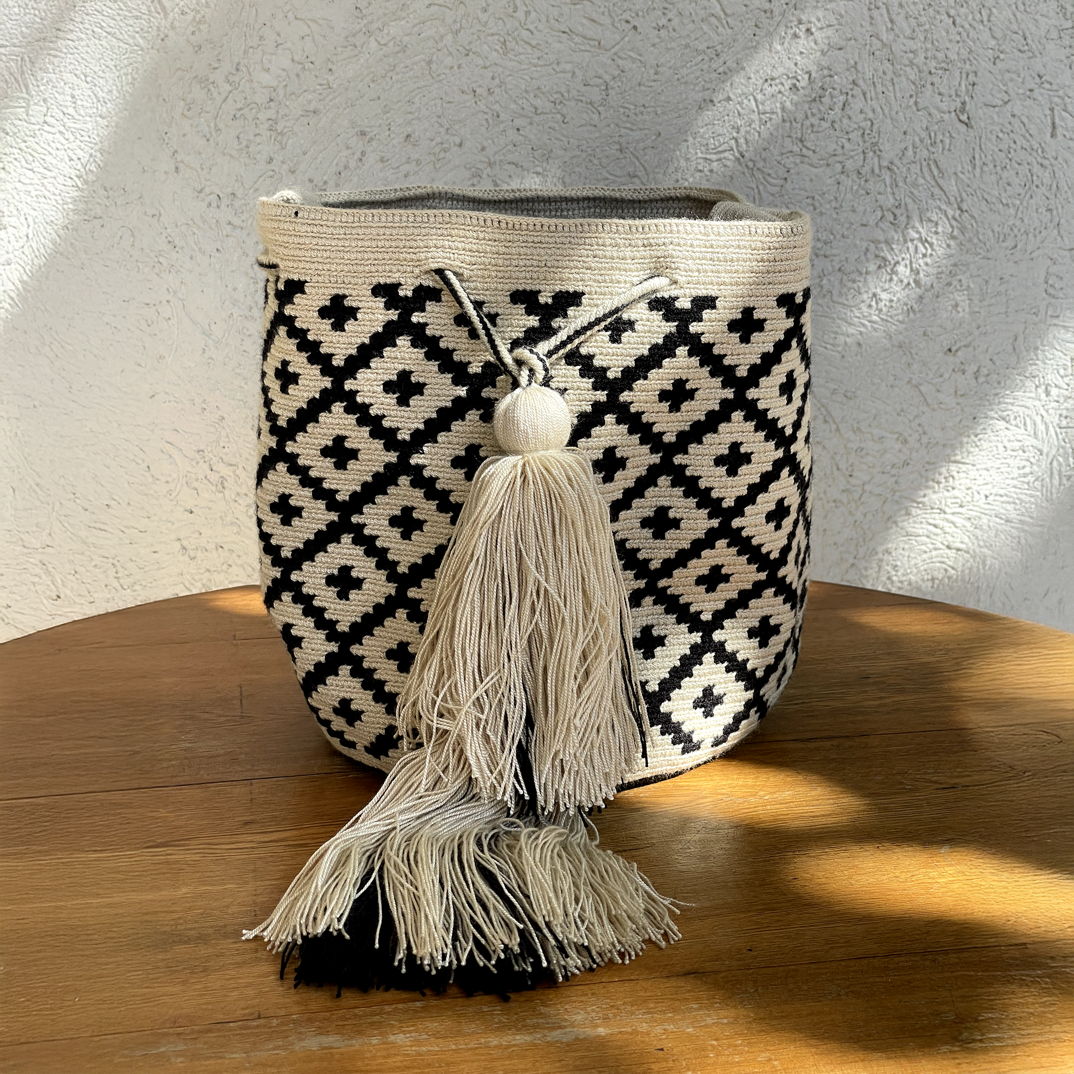 Mochila Wayúu con diseño de rombos negros sobre fondo beige, tejida a mano con técnica de crochet y decorada con una borla grande.