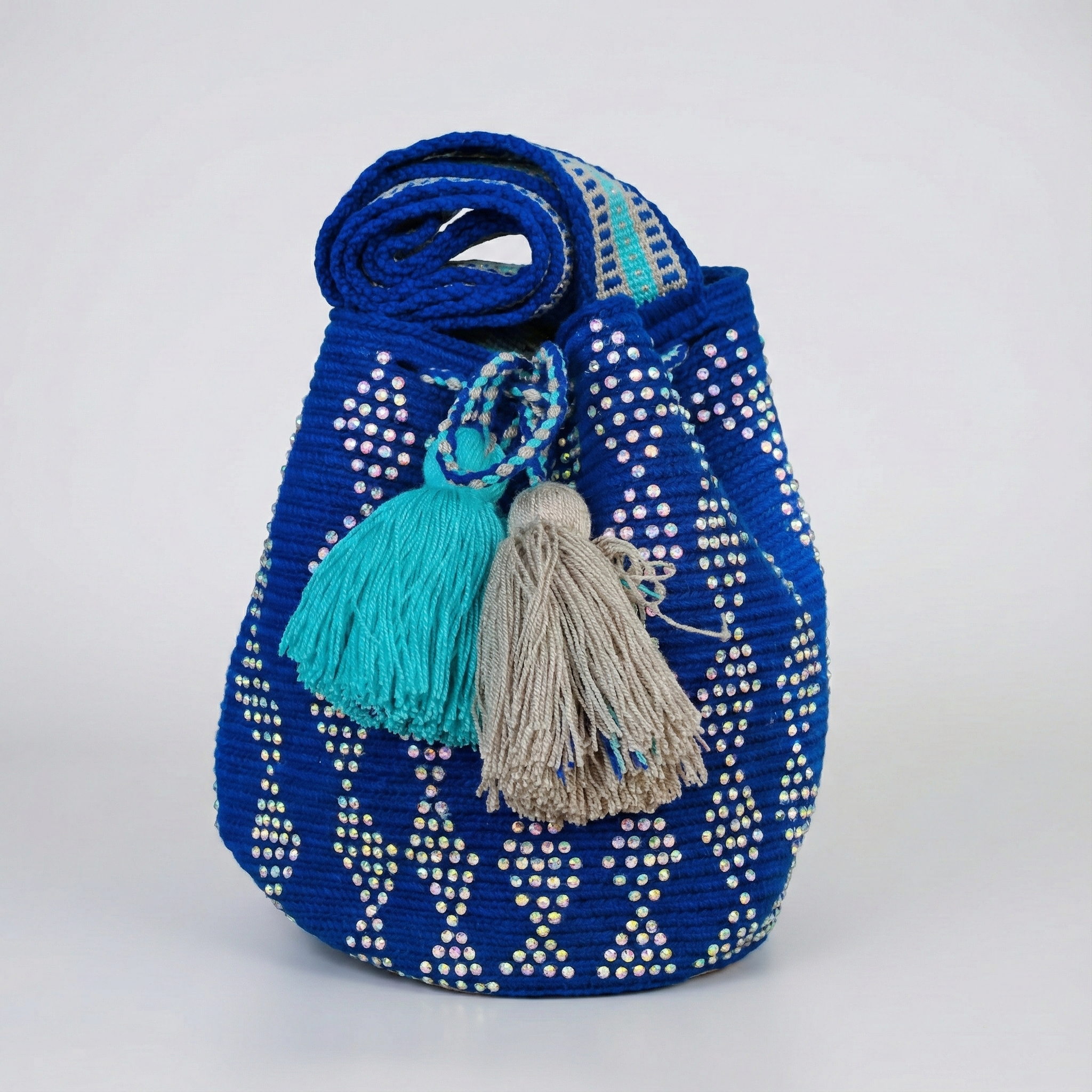 Mochila Wayúu artesanal de color azul rey decorada con cristales brillantes en patrones geométricos, con gasa tejida y dos borlas grandes en colores turquesa y beige.