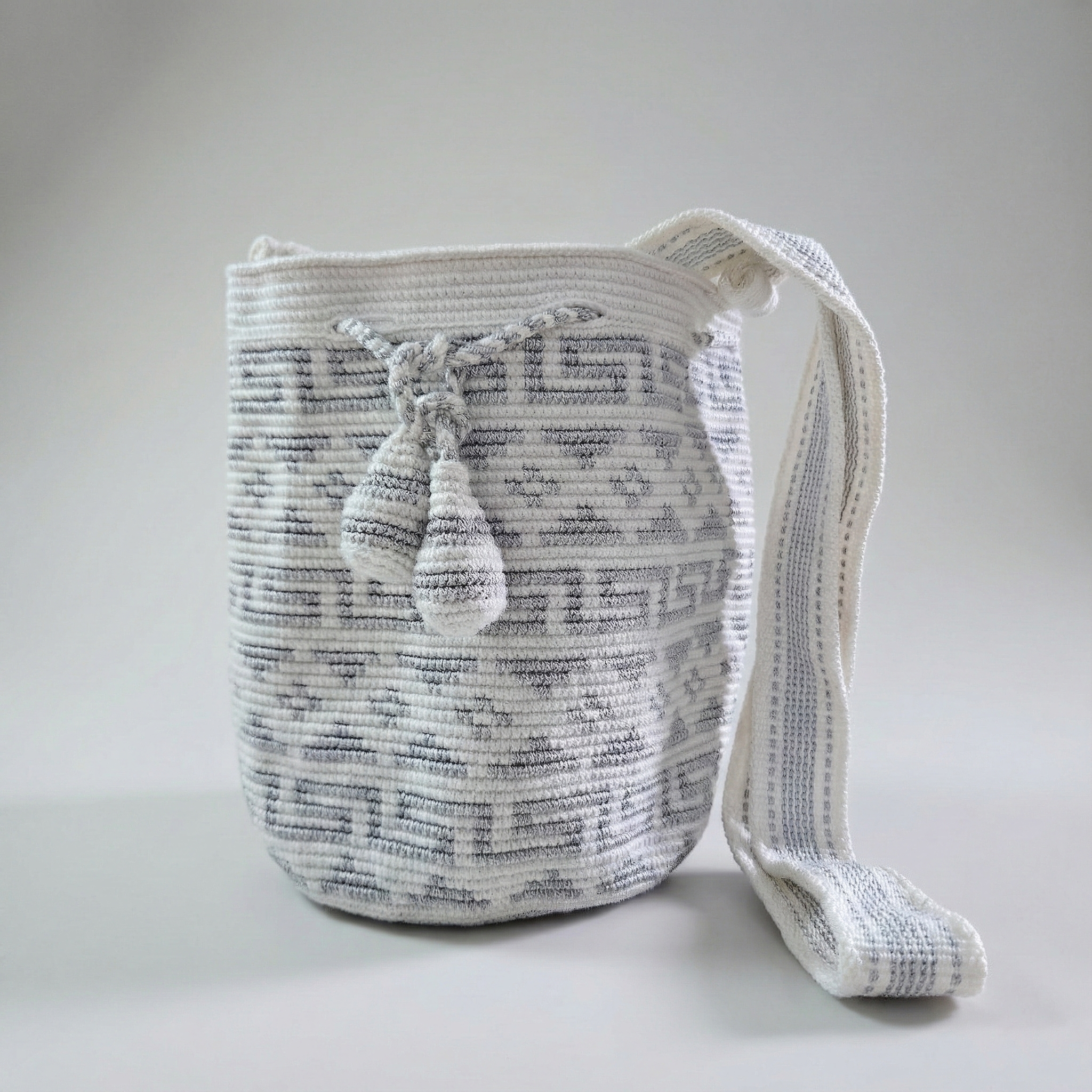 Mochila Wayúu artesanal en color blanco con diseños geométricos Kanaas en color gris, cordón de ajuste con borlas y correa ancha tejida.