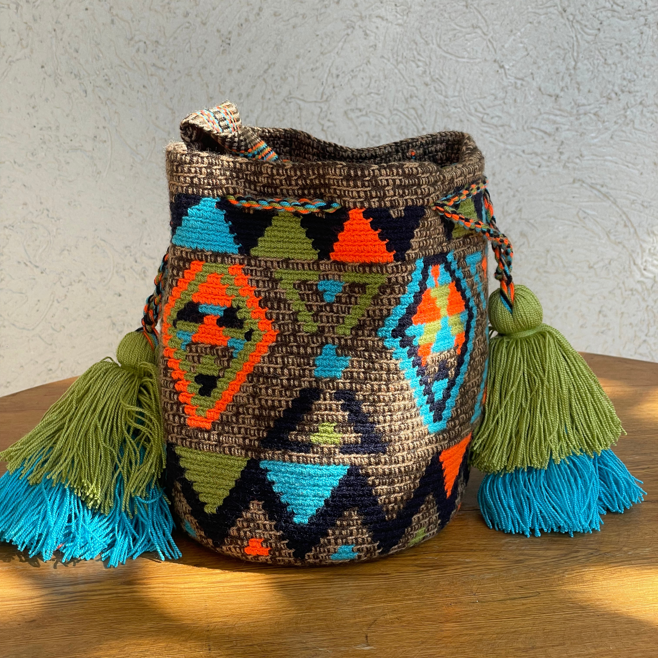 Mochila Wayúu de color café con diseños geométricos en naranja, azul turquesa y verde, con borlas grandes decorativas y correa tejida.