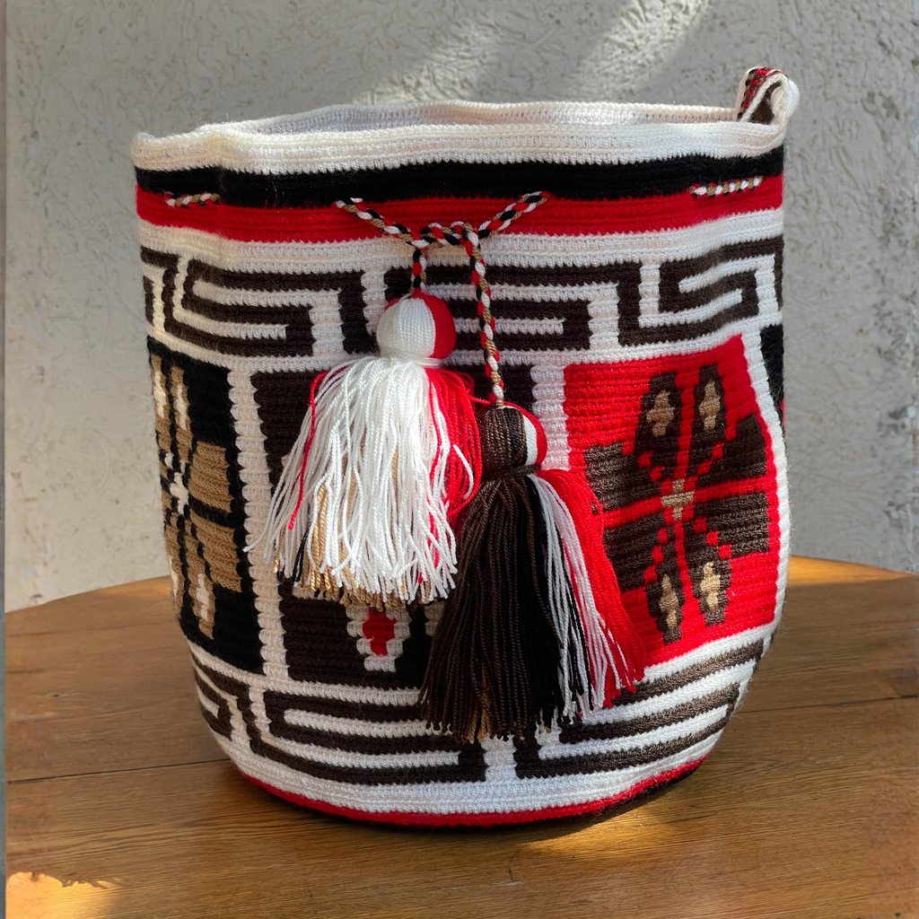 Mochila Wayúu tejida a mano con patrones geométricos Kanaas en colores blanco, rojo y café, con dos borlas grandes frontales.