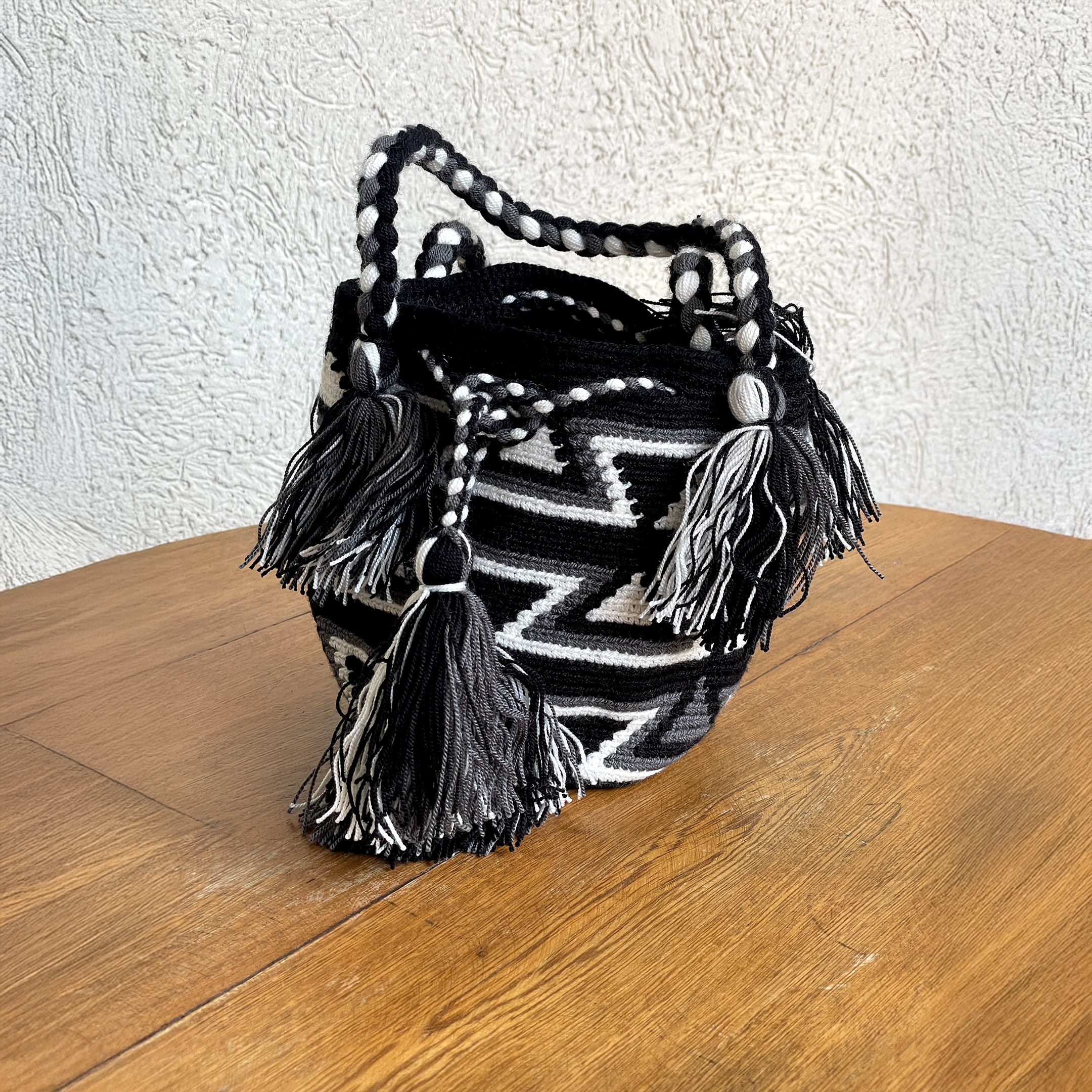 Mochila Wayúu con diseño de figuras geométricas Kanaas en colores negro, gris y blanco, con borlas grandes y gasa trenzada sobre fondo de madera.