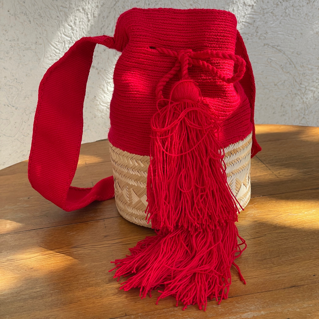 Mochila Wayúu artesanal roja con base de fibra natural tejida, cordón de ajuste y borla grande roja sobre fondo de madera.