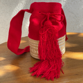 Mochila Wayúu artesanal roja con base de fibra natural tejida, cordón de ajuste y borla grande roja sobre fondo de madera.