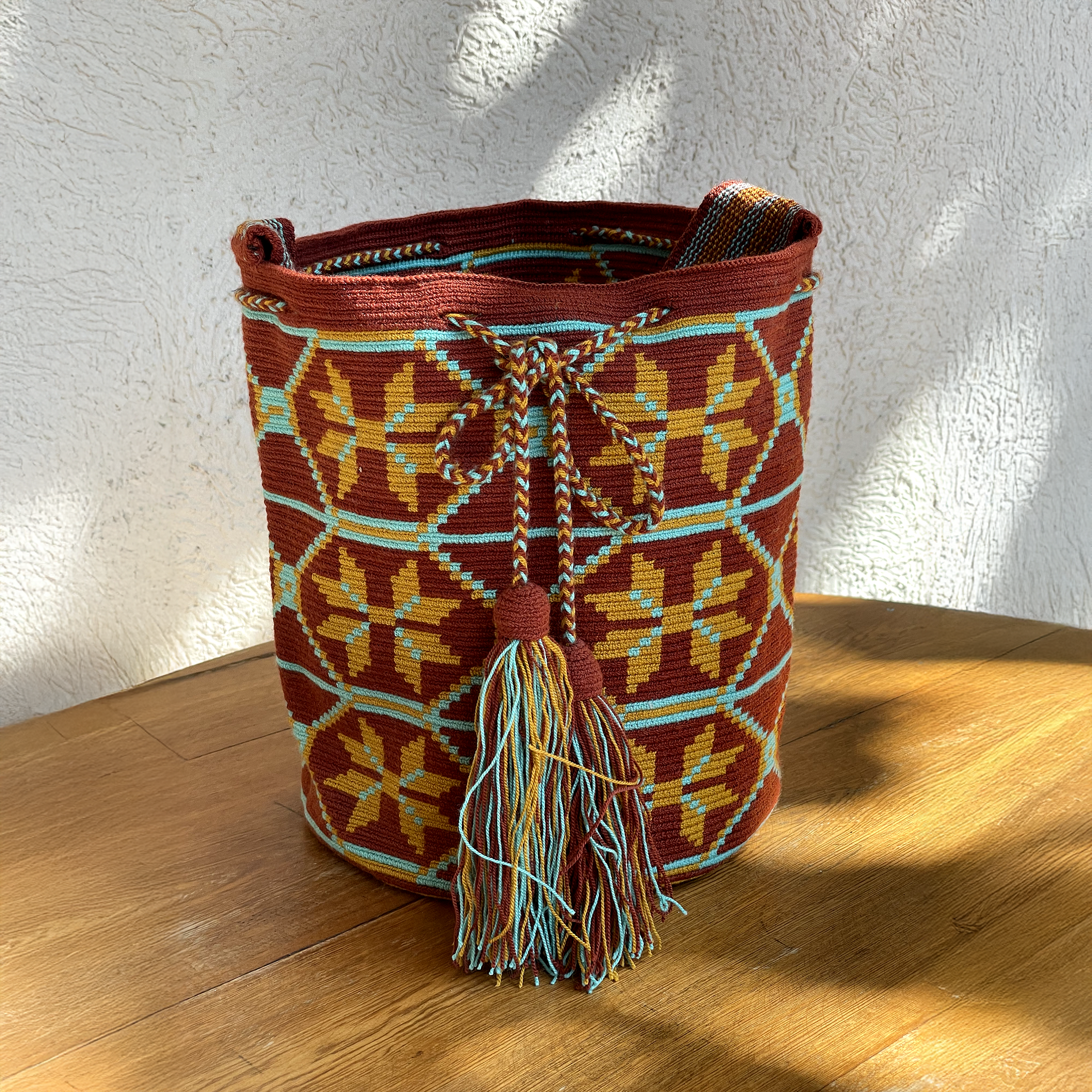 Mochila Wayúu tejida a mano en color terracota con figuras geométricas amarillas y turquesas, cordón trenzado y borlas grandes sobre mesa de madera.