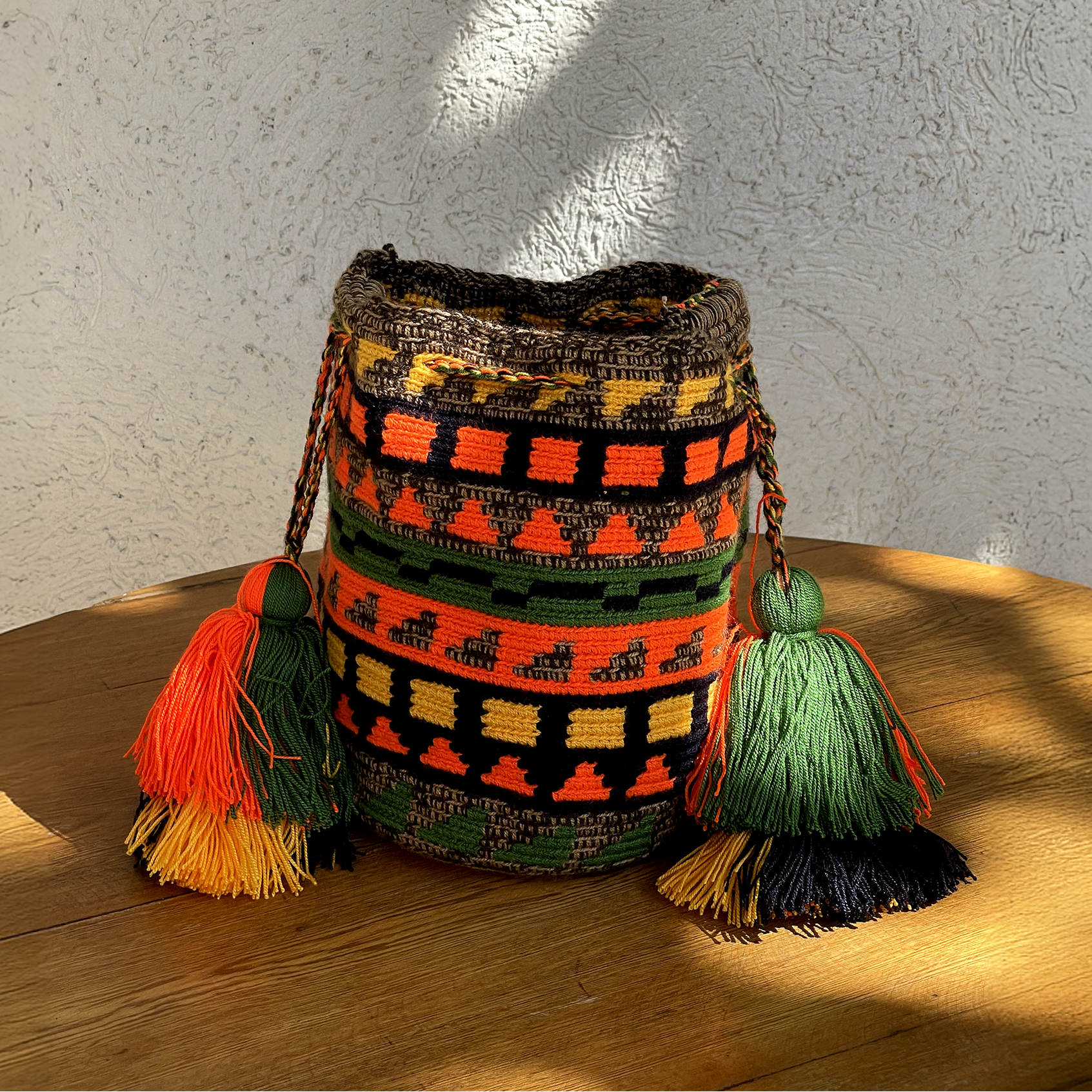 Mochila Wayúu tejida a mano con patrones de franjas geométricas en naranja, amarillo, verde y café con borlas grandes.