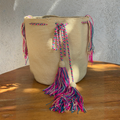 Mochila Wayúu de color beige con cordón trenzado y borlas grandes en colores fucsia y azul, sobre una mesa de madera.