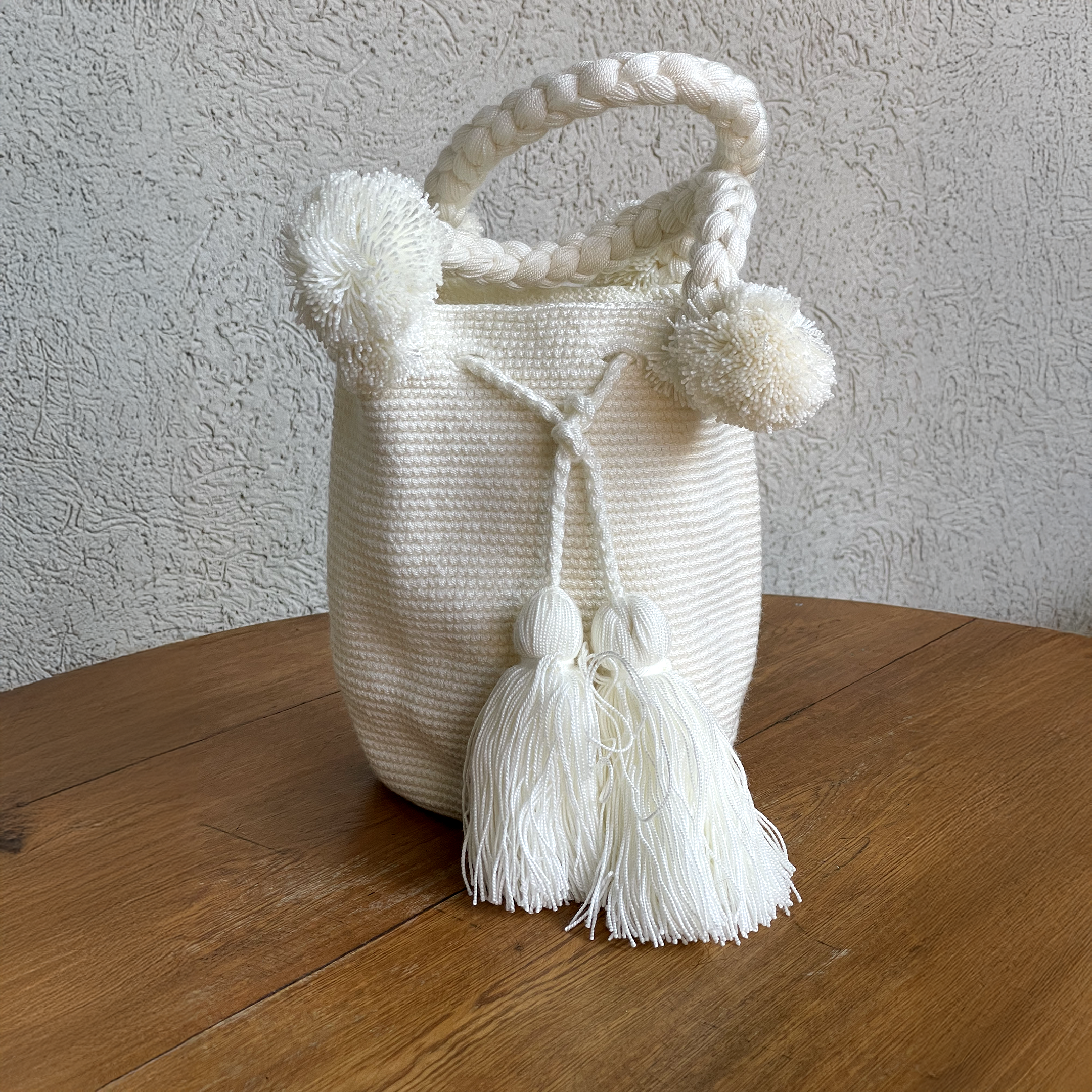 Mochila Wayúu artesanal unicolor en color blanco crudo, con asa trenzada gruesa, pompones decorativos y borlas grandes frontales sobre una mesa de madera.