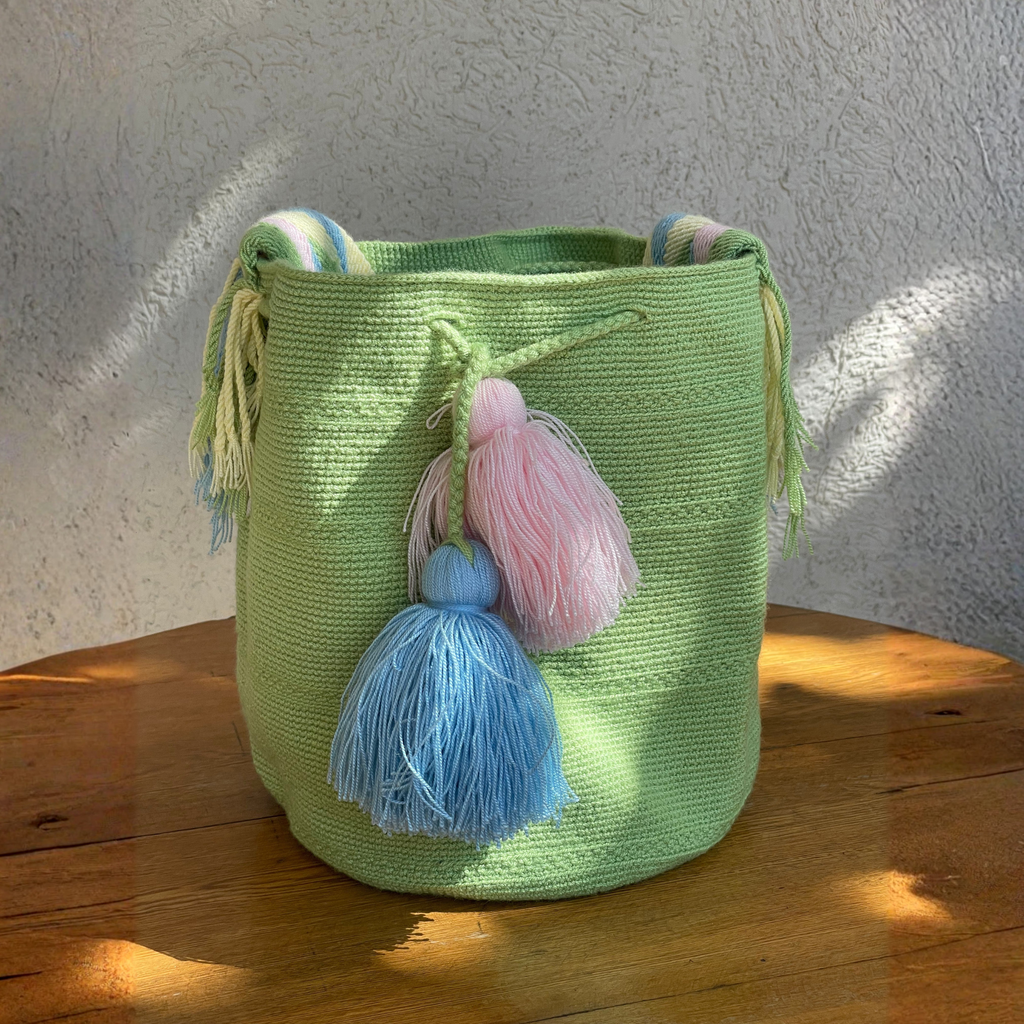 Mochila Wayúu de color verde lima tejida a mano con grandes borlas en azul y rosa pastel sobre una mesa de madera.