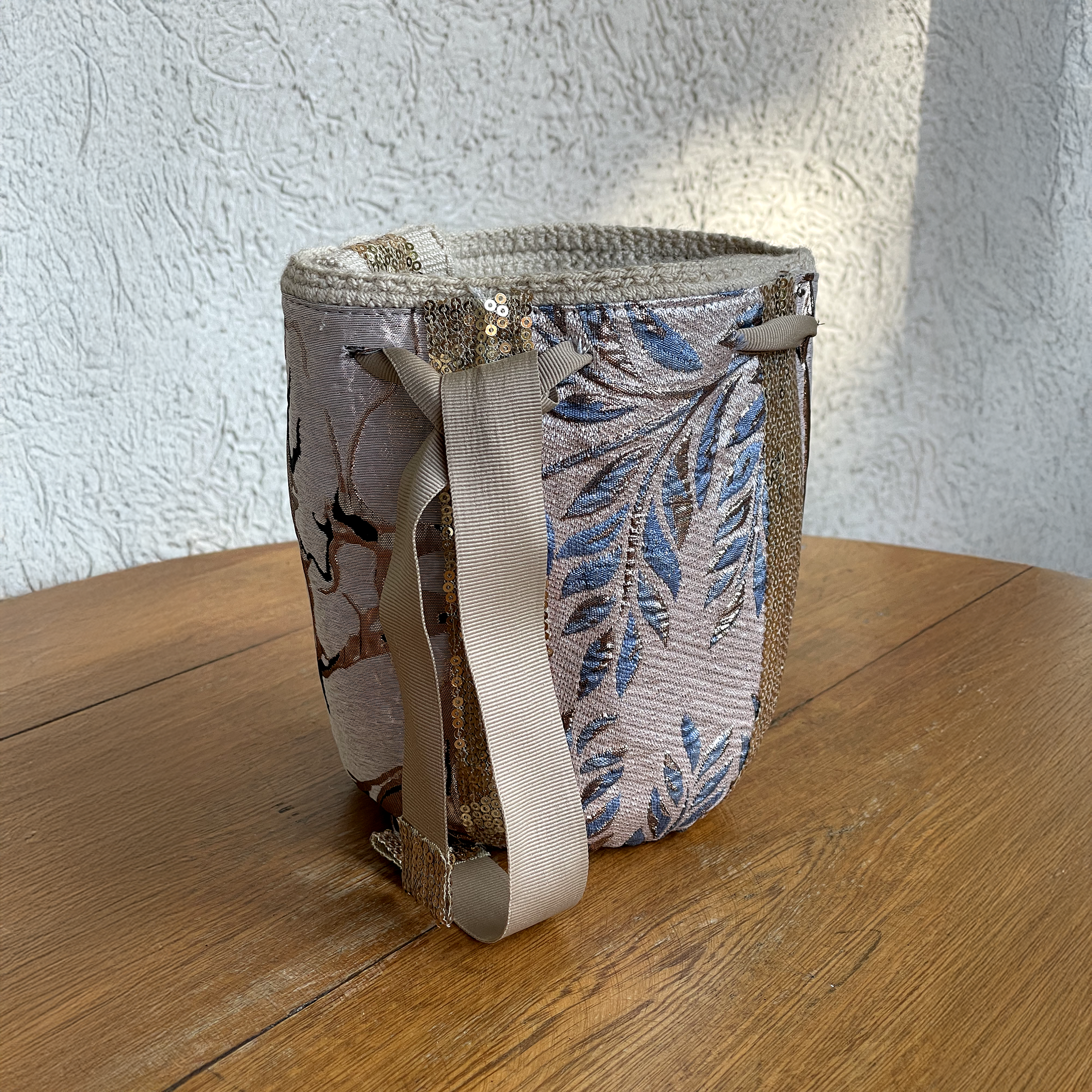Mochila Wayúu de gala con diseño floral azul y bronce, adornada con bandas verticales de lentejuelas doradas y cierre de cinta color arena.