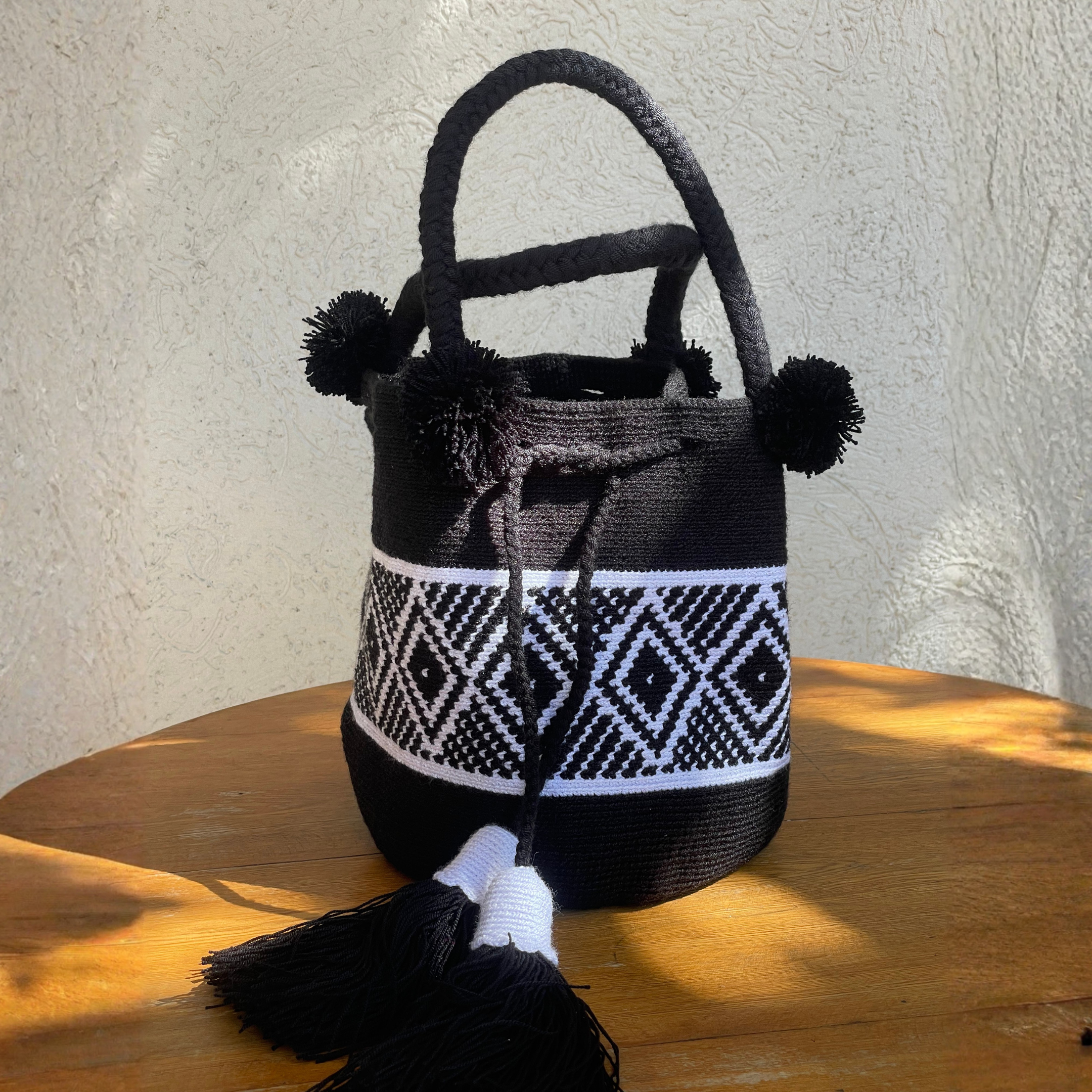 Mochila Wayúu de mano color negro con diseño de figuras geométricas blancas (Kanaas), doble asa trenzada y borlas grandes decorativas.