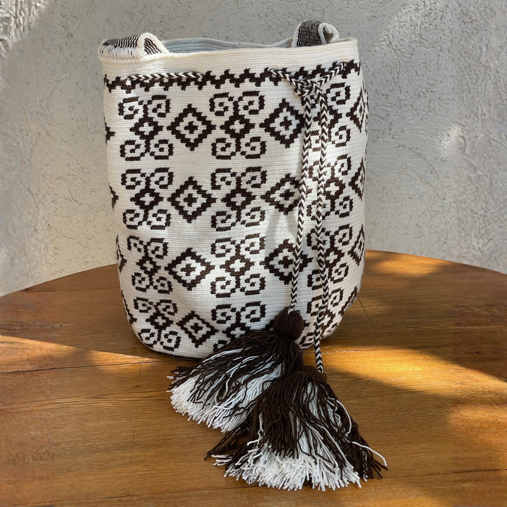Mochila Wayuu artesanal en color blanco con patrones geométricos café, cordón trenzado y borlas grandes.