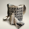 Mochila Wayuu artesanal con diseño geométrico de laberinto en blanco y negro, con dos borlas grandes y gaza ancha tejida.