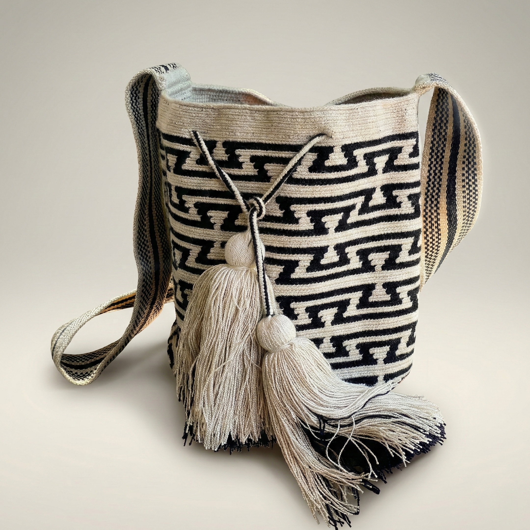 Mochila Wayuu artesanal con diseño geométrico de laberinto en blanco y negro, con dos borlas grandes y gaza ancha tejida.