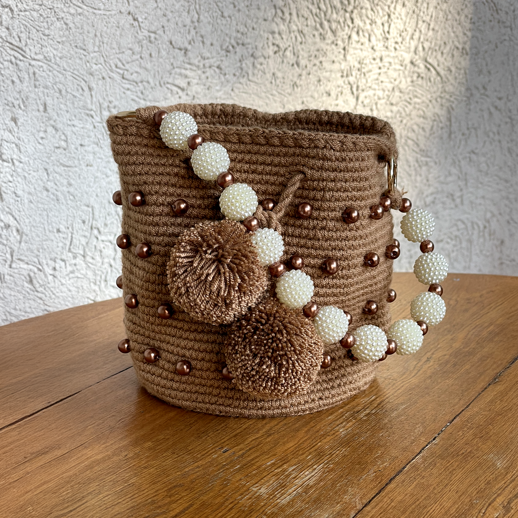 Mochila Wayuu de color café con apliques de perlas bronce en el cuerpo, asa de perlas blancas y borlas grandes decorativas sobre una superficie de madera.