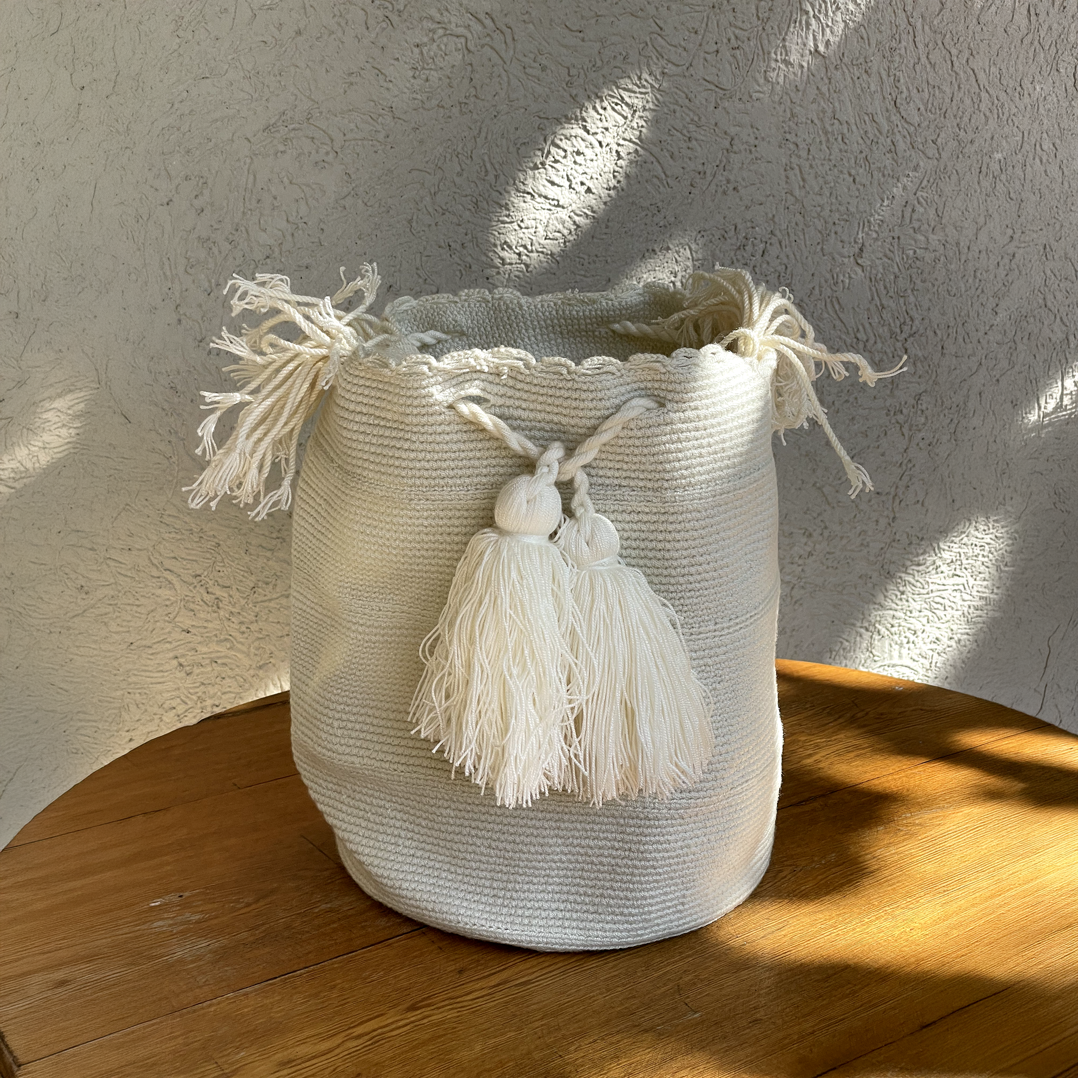 Mochila Wayúu unicolor en tono blanco con tejido de punto artesanal, cordón trenzado y borlas grandes decorativas sobre una mesa de madera.