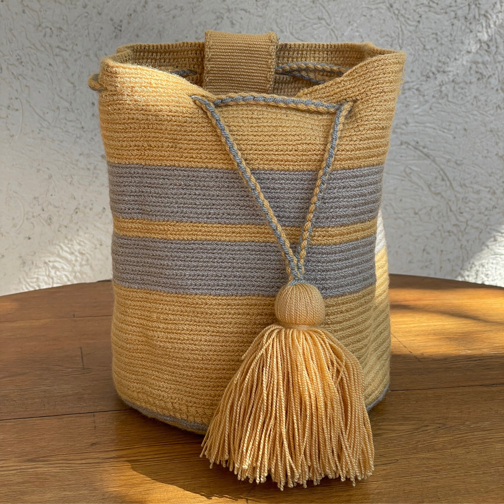 Mochila Wayuu tejida a mano con diseño de rayas horizontales en color amarillo y gris, incluye cierre de cordón trenzado y borla decorativa amarilla.