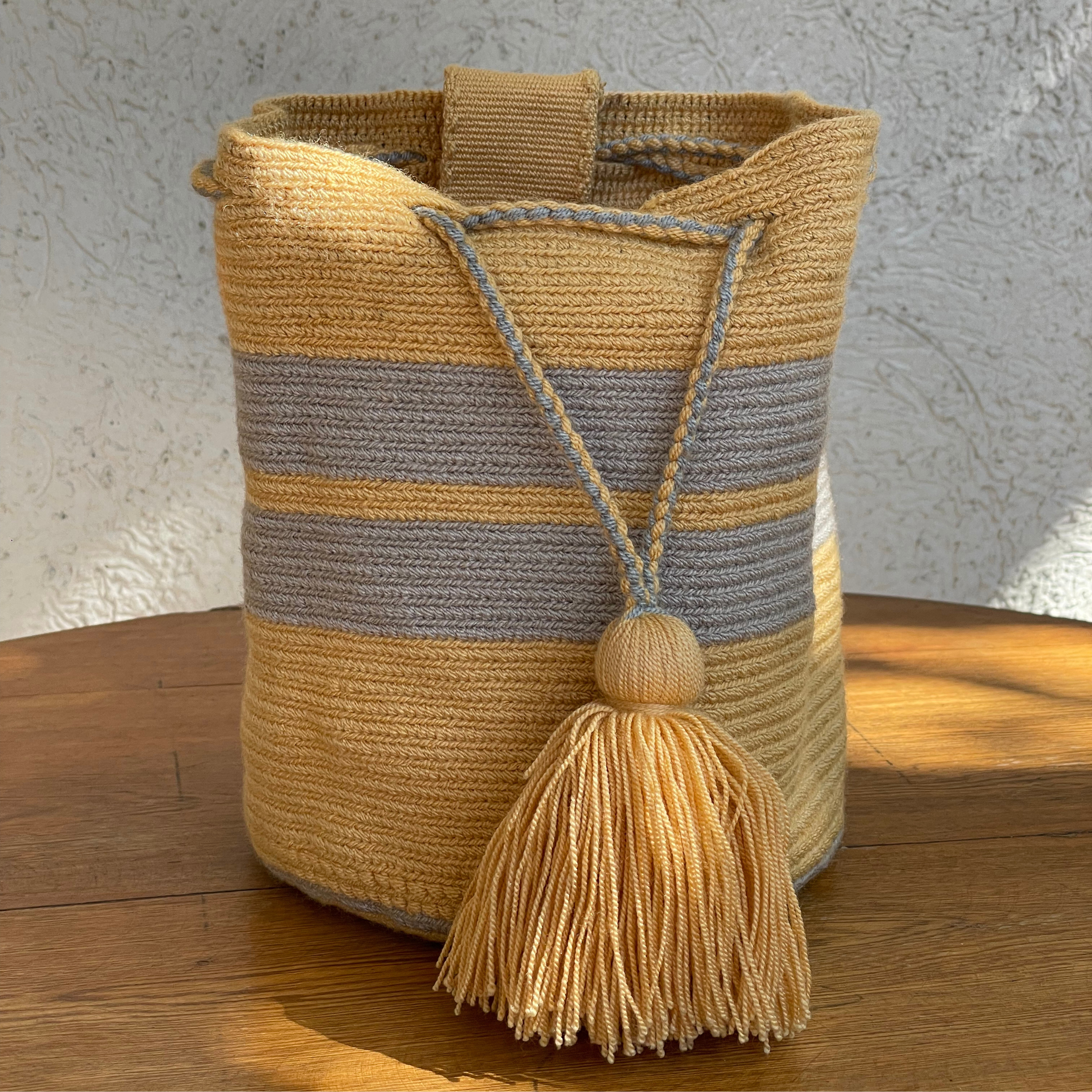 Mochila Wayuu tejida a mano con diseño de rayas horizontales en color amarillo y gris, incluye cierre de cordón trenzado y borla decorativa amarilla.